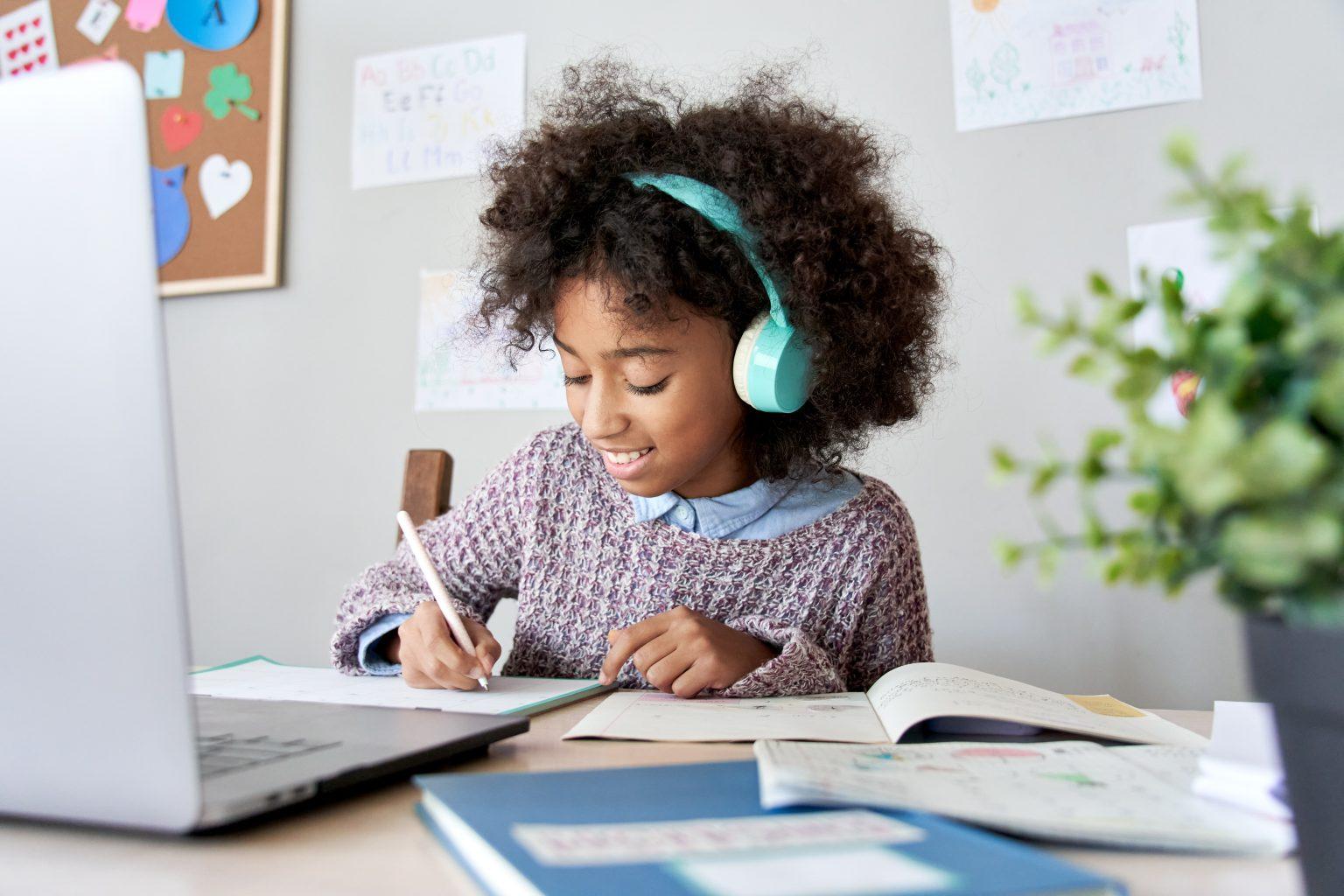 A young girl wearing headphones is focused on writing while seated in front of a laptop.