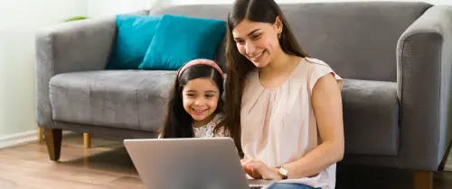 A woman and a child are seated on the floor, engaged with a laptop in a cozy indoor setting.