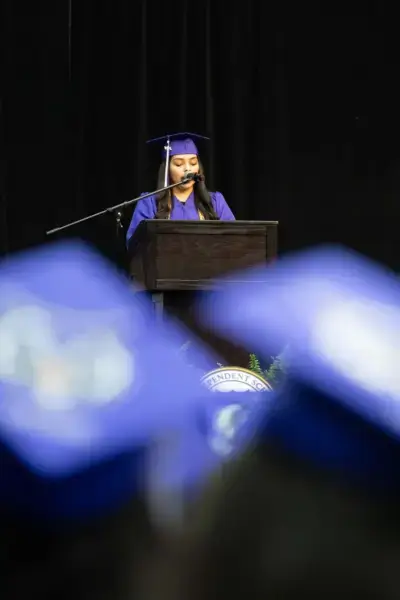 A woman in a purple graduation gown and cap stands at a podium, speaking into a microphone. Blue caps of seated graduates blur in the foreground. The atmosphere is formal and celebratory.