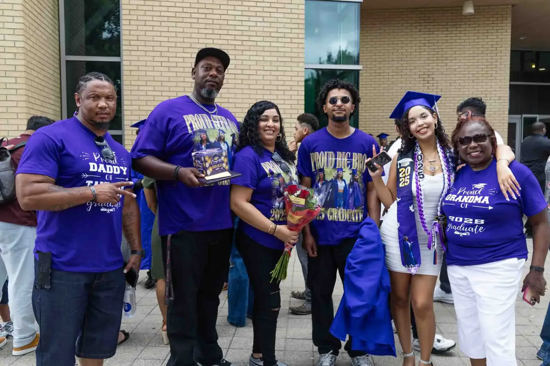 A joyful family celebrating graduation. Six people stand together outside, wearing shirts with supportive messages. The graduate, in cap and gown, smiles brightly.