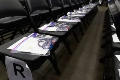 Rows of black chairs with packets on each seat, including a booklet and a colorful cord. A paper labeled "R" marks a row. The setting is calm and organized.