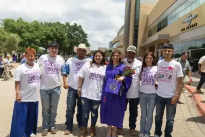 A group of eight people pose outdoors. A woman in a purple graduation gown holds flowers, surrounded by supportive family members wearing custom t-shirts. The atmosphere is celebratory.