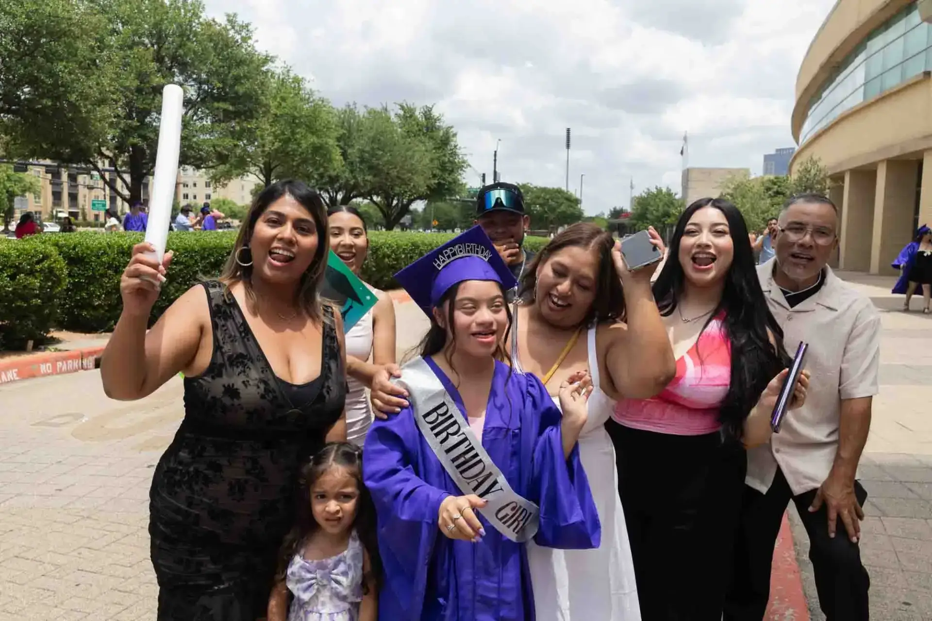 Happy graduate in a purple cap and gown, wearing a "Birthday Girl" sash, surrounded by joyful family holding a diploma. Outdoor celebration, sunny day.